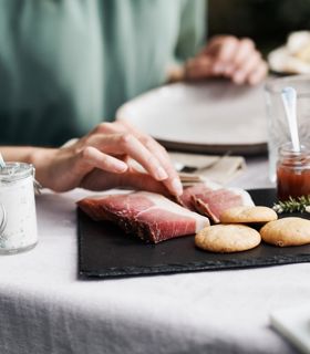Una persona prende una fetta di speck da un tagliere con specialità altoatesine come formaggio, pane, quark alle erbe e marmellata.