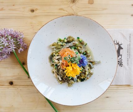 Piatto con pasta, verdure, salmone e fiori edibili su un tavolo di legno chiaro con un fiore.